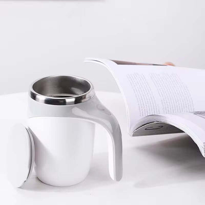 Magnetic self-stirring mug in white with a stainless steel interior, next to an open magazine on a table.